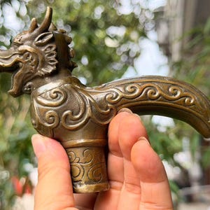 May include: A bronze-colored metal cane handle shaped like a dragon head with an open mouth and detailed swirling patterns along the neck and curved grip. The object features a cylindrical base and a textured finish with dark patina accents.