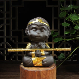 May include: A dark brown ceramic monkey figurine in a seated meditation pose, holding a gold-colored staff. The monkey has closed eyes and gold accents on the head and clothing. The figurine is displayed on a wooden base.