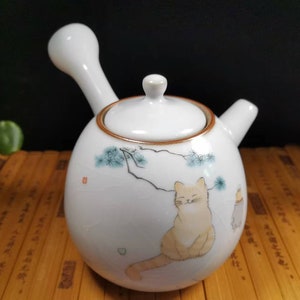 Könnte beinhalten: Weiße Keramik-Teekanne mit einer Katzenillustration. Die Teekanne hat einen gebogenen Griff, einen kleinen Ausguss und einen Deckel. Die Katze ist in sitzender Position mit geschlossenen Augen dargestellt. Die Teekanne hat einen braunen Rand und einen Ast mit blauen Blättern.
