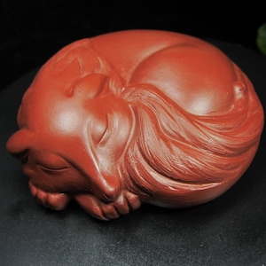 Może przedstawiać: Ceglastobrązowa rzeźba ceramiczna śpiącego lisa, zwiniętego w kulkę. Sierść lisa jest szczegółowa z wyrzeźbionymi liniami, a jego oczy są zamknięte. Rzeźba znajduje się na czarnej powierzchni.