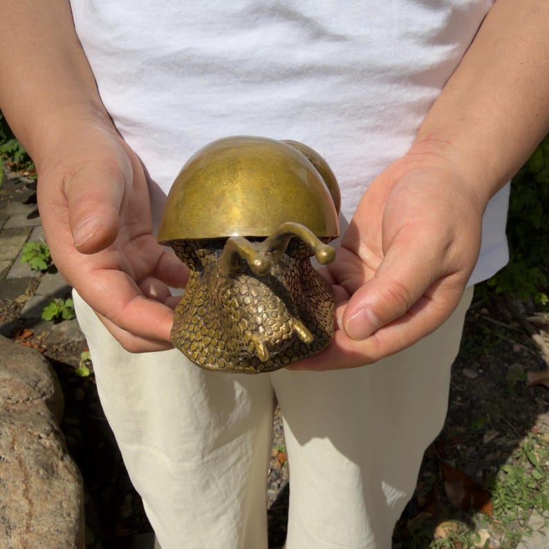 May include: A brass snail figurine featuring a smooth, polished, dome-shaped shell and a textured, bumpy body. The snail has two prominent eye stalks extending forward from its head, with a metallic gold finish throughout the entire object.