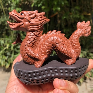Puede incluir: Figura detallada de dragón de terracota con la boca abierta, escamas y cola en forma de aleta. El dragón está montado sobre una base oscura y texturizada. La escultura es de color marrón rojizo.