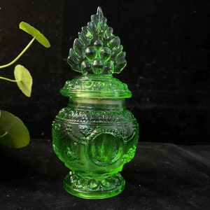 Könnte beinhalten: Ein grünes Glasgefäß mit dekorativem Deckel. Das Gefäß hat ein verziertes Design mit detailliertem Sockel und einem Deckel, der von einem blattartigen Design gekrönt wird. Das Gefäß besteht aus durchscheinendem grünem Glas.