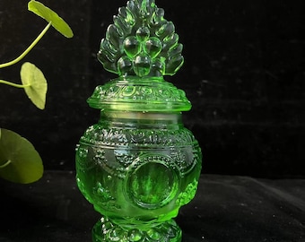 May include: A green glass jar with a decorative lid. The jar features an ornate design with a detailed base and a lid topped with a leaf-like design. The jar is made of translucent green glass.