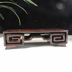 Op de afbeelding: Een donkerbruine houten displaystandaard met een rechthoekige bovenkant en een decoratief, gesneden ontwerp. De standaard heeft een centrale opening en opgerolde details aan de zijkanten. De standaard is ongeveer 20 cm lang.