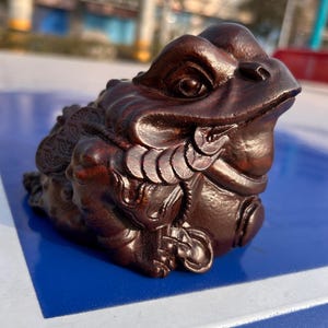 May include: A detailed, wooden sculpture of a frog, featuring intricate carvings and a rich, dark brown finish. The frog is depicted with coins, suggesting wealth and prosperity. The sculpture is set against a blue surface.