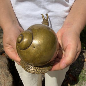 May include: A polished brass snail figurine featuring a smooth, rounded shell and a textured base. The snail has two upright antennae and a detailed, pebbled surface pattern on its lower body, showcasing a metallic gold-toned finish throughout.