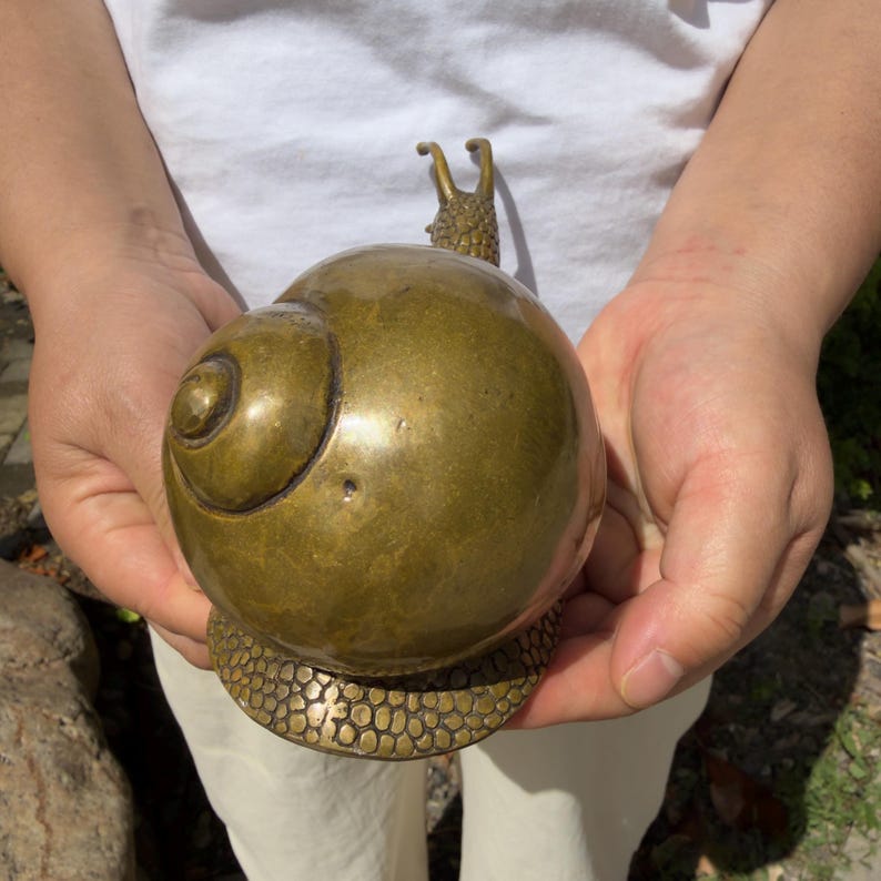May include: A polished brass snail figurine featuring a smooth, rounded shell and a textured base. The snail has two upright antennae and a detailed, pebbled surface pattern on its lower body, showcasing a metallic gold-toned finish throughout.