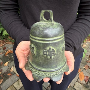 Könnte beinhalten: Eine dunkelgrüne, verwitterte Bronzeglocke mit einer abgerundeten oberen Öse und einem ausgestellten, gewellten unteren Rand. Die Oberfläche zeigt erhabene, geprägte Schriftzeichen und ein dekoratives Rankenmuster an der Basis.