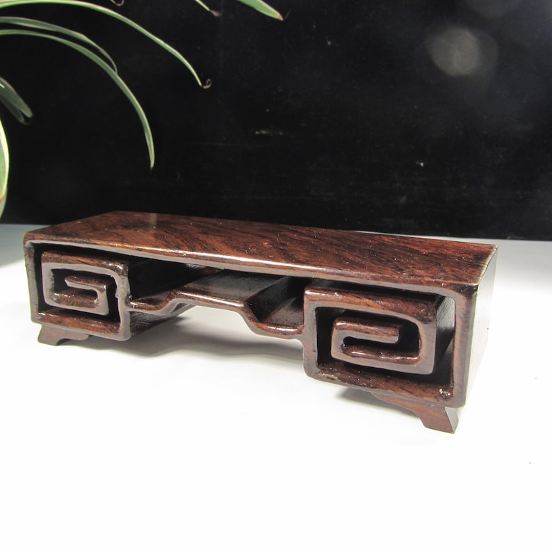 Op de afbeelding: Een rechthoekige, donkerbruine houten displaystandaard met een glanzende afwerking. De standaard heeft een verzonken midden en decoratieve, wervelende uitsparingen aan elk uiteinde. De standaard is ongeveer 25 cm lang en 8 cm breed.