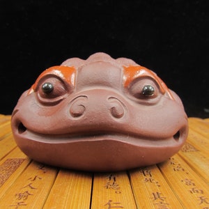 May include: A brown ceramic frog figurine with orange accents above the eyes. The frog has black bead eyes and a smiling mouth. The figurine is sitting on a bamboo surface.