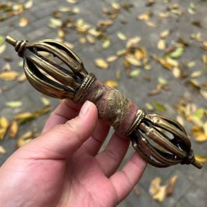 May include: A brass vajra ritual object featuring two symmetrical, cage-like ends with curved prongs and a central handle wrapped in a textured, weathered reddish-brown fabric. The metal has a tarnished, antique finish with visible oxidation.
