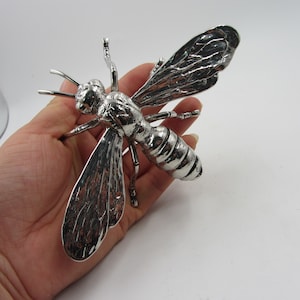 May include: A silver-colored metal bee sculpture with detailed wings, antennae, and body segments. The bee is shown in a close-up view, highlighting its intricate design and metallic sheen. The sculpture is a decorative item.