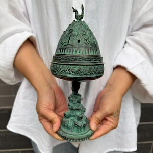 May include: A dark green, oxidized metal incense burner featuring an intricate, conical lid with a small finial at the top. The body is decorated with embossed patterns and rests on a sculpted, textured base with a wide, circular foot.