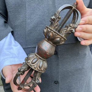 Può includere: Un oggetto rituale vajra in metallo scuro e usurato, caratterizzato da un'impugnatura sferica centrale affiancata da due basi a petali di loto. Ogni estremità è composta da quattro punte ricurve che convergono, con tracce di vernice rossa.