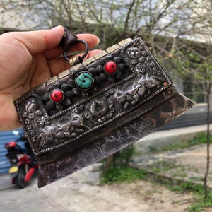 Pode incluir: Uma faca antiga prateada com intrincadas gravuras florais e de animais. A pega apresenta pedras vermelhas e turquesas. Uma presilha de couro e um anel de metal estão presos no topo. A lâmina tem uma aparência desgastada e envelhecida.