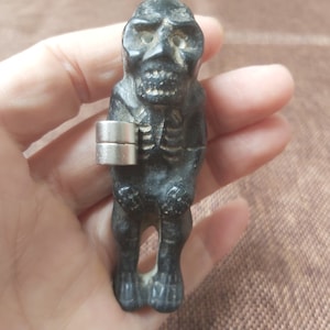 May include: A small black figurine of a skeleton with a skull head. The figurine is wearing a silver ring on its arm.