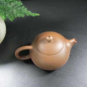 Könnte beinhalten: Eine kleine, braune Keramik-Teekanne mit rundem Körper, gebogenem Henkel und Ausguss. Der Deckel hat einen kleinen Knopf und ein Loch. Die Teekanne ist zum Aufbrühen von Tee bestimmt.