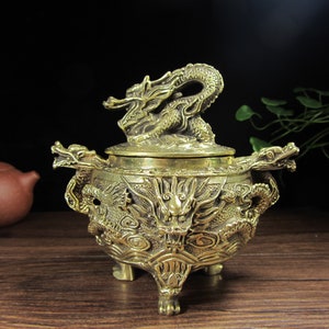May include: A brass incense burner with intricate dragon detailing. The burner features a dragon on the lid and handles, with additional dragon and cloud motifs on the body. It has three clawed feet and a round shape.