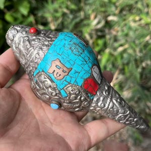 Peut inclure: Coquillage en argent orné avec incrustations turquoise et accents rouges et bleus. Le coquillage présente des gravures complexes et est tenu dans une main. Un symbole couleur cuivre est serti dans la turquoise.