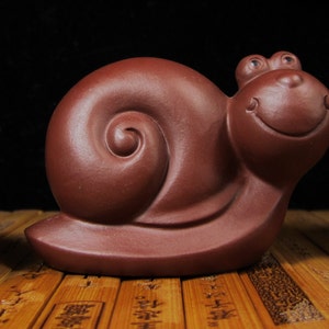 Könnte beinhalten: Eine skurrile, rotbraune Keramik-Schneckenfigur mit einem lächelnden Gesicht und großen, ausdrucksstarken Augen. Das Schneckenhaus hat ein Spiraldesign, und die Figur hat eine glatte, matte Oberfläche. Ein dekorativer Artikel.