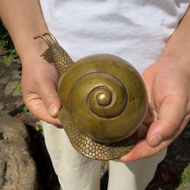 May include: A brass snail figurine featuring a smooth, rounded shell with a prominent spiral pattern and a textured, scale-like surface on the head and foot. The metallic object has a warm, golden-brown finish and two distinct eye stalks.
