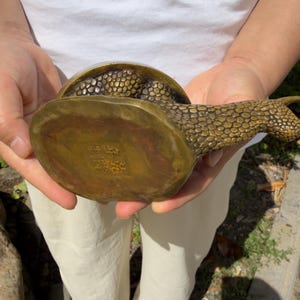 May include: A brass snail figurine with a textured, pebbled surface on its shell and body. The underside of the snail features a square stamp with four Chinese characters engraved into the metal, showing a metallic gold and bronze finish.