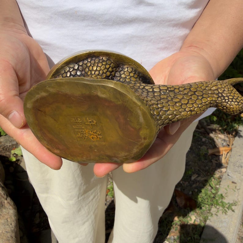 May include: A brass snail figurine with a textured, pebbled surface on its shell and body. The underside of the snail features a square stamp with four Chinese characters engraved into the metal, showing a metallic gold and bronze finish.