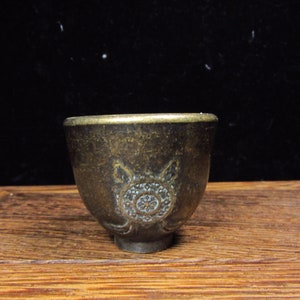Può includere: Una piccola tazza antica color bronzo con un disegno dettagliato di tartaruga sul davanti. La tazza ha una forma arrotondata con un bordo leggermente svasato. Il disegno della tartaruga è centrato e intricato.
