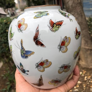 Puede incluir: Jarrón de cerámica blanca adornado con ilustraciones de mariposas coloridas. Las mariposas presentan una variedad de colores, incluyendo rojo, amarillo, azul y verde. El jarrón tiene forma redondeada y un borde marrón.