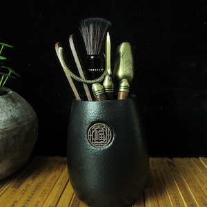 May include: A black, textured ceramic tea tool holder with a bronze emblem. The holder contains a set of tea ceremony tools, including a brush, tongs, and a scoop. The tools have bronze and wooden handles.