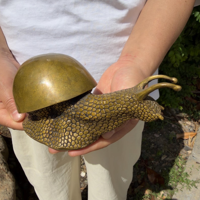 May include: A brass snail figurine featuring a smooth, rounded, polished shell and a textured, bumpy body with two extended eye stalks. The metal has a warm, golden-brown patina with visible variations in tone across the surface of the sculpture.