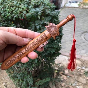 Puede incluir: Una espada decorativa de madera con intrincadas tallas y una borla roja. La espada presenta un mango con un símbolo Yin Yang y el número "1208" grabado en la hoja. La hoja tiene tallas detalladas de flores y dragones.