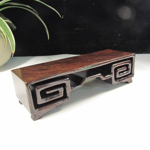 Könnte beinhalten: Ein rechteckiger, dunkelbrauner Holz-Präsentationsständer mit glänzender Oberfläche. Der Ständer hat eine eingelassene Ablage und dekorative, geschnitzte Schnörkel an den Enden. Er steht auf kleinen Füßen und zeigt ein klassisches Design.