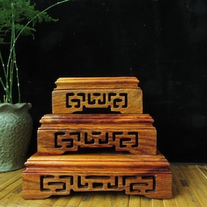 May include: Three wooden display stands of different sizes are stacked. Each stand is rectangular with a decorative cutout design. The wood grain is visible, and the stands have a warm, reddish-brown hue. A green plant in a pot is visible in the background.