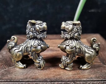 May include: Two antique brass lion statues. Each lion is in a seated position, holding a ball under its paw. The lions have detailed manes and tails, with an aged, weathered appearance. The statues are displayed on a wooden surface.
