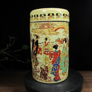 Puede incluir: Un recipiente cilíndrico de cerámica con tapa, que presenta una escena pintada de figuras con vestimenta tradicional japonesa. El recipiente tiene una base de color crema con un borde decorativo y una base negra.