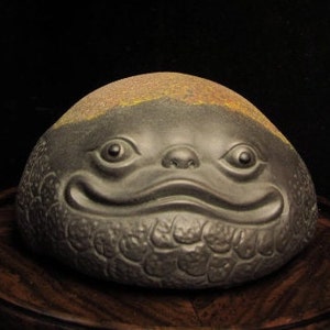 Puede incluir: Una escultura redondeada de color gris oscuro con una cara sonriente. La escultura tiene una superficie texturizada y una banda de material dorado en la parte superior. Los ojos, la nariz y la boca están claramente definidos, lo que le da a la escultura una apariencia caprichosa. Se encuentra sobre una superficie de madera oscura.