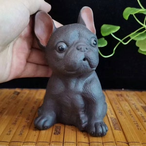 May include: A dark brown ceramic figurine of a French Bulldog. The dog has large, expressive eyes, a wrinkled face, and large, upright ears. The figurine is sitting on a bamboo surface.