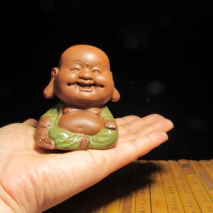 May include: A small, smiling Buddha figurine. The ceramic statue is brown with a green robe. The Buddha is sitting with its legs crossed and has large earlobes. The figurine is held in a person's hand against a black background.