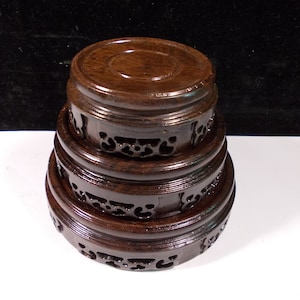 Puede incluir: Una pila de tres soportes de exhibición de madera marrón oscuro. Cada soporte es circular con una parte superior plana y patrones decorativos recortados en los lados. Los soportes son de diferentes tamaños, creando un efecto escalonado.