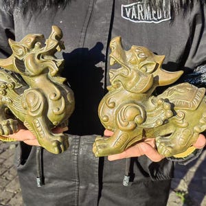 May include: Two bronze-colored mythical creature statues, each with a lion-like body, open mouth, and intricate details. The statues are held up, showcasing their detailed craftsmanship and design. The statues are likely for home decor.