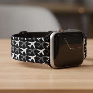 May include: A silver smartwatch with a black screen and a black and white airplane patterned band. The watch is displayed on a light brown wooden surface.