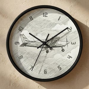 May include: Round wall clock with a black frame. The clock face features a pencil sketch of a small airplane in flight against a cloudy sky. Black hands indicate the time, and the numbers are in a simple, modern font.