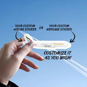 Op de afbeelding: Een witte vliegtuigsticker met de tekst "Emirates" en de woorden "YOUR CUSTOM AIRLINE STICKER OR YOUR CUSTOM AIRPLANE STICKER" en "CUSTOMIZE IT AS YOU WISH!" tegen een blauwe lucht.