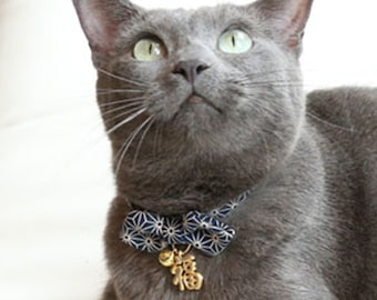 May include: A gray cat wearing a blue and white patterned bow tie with a gold charm. The cat is looking up and has green eyes.