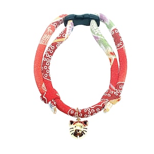 Puede incluir: Collar de gato rojo y dorado con un dije en forma de gato. El collar está hecho de tela con un patrón japonés tradicional.