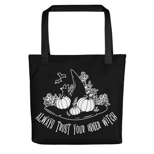 Può includere: Borsa tote nera con un'illustrazione bianca di un cappello da strega, zucche, un gatto nero e pipistrelli. Il testo "ALWAYS TRUST YOUR INNER WITCH" è stampato a forma di arco. La borsa ha manici neri.