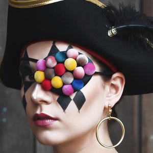 May include: A woman wearing a black pirate hat with gold trim and a black eye patch decorated with colourful pom-poms. Her face is painted with harlequin diamonds and white accents, and she wears gold hoop earrings.