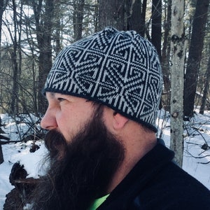Può includere: Un cappello di lana nero e bianco con un motivo geometrico. Il cappello è indossato da una persona con una lunga barba in un bosco innevato.